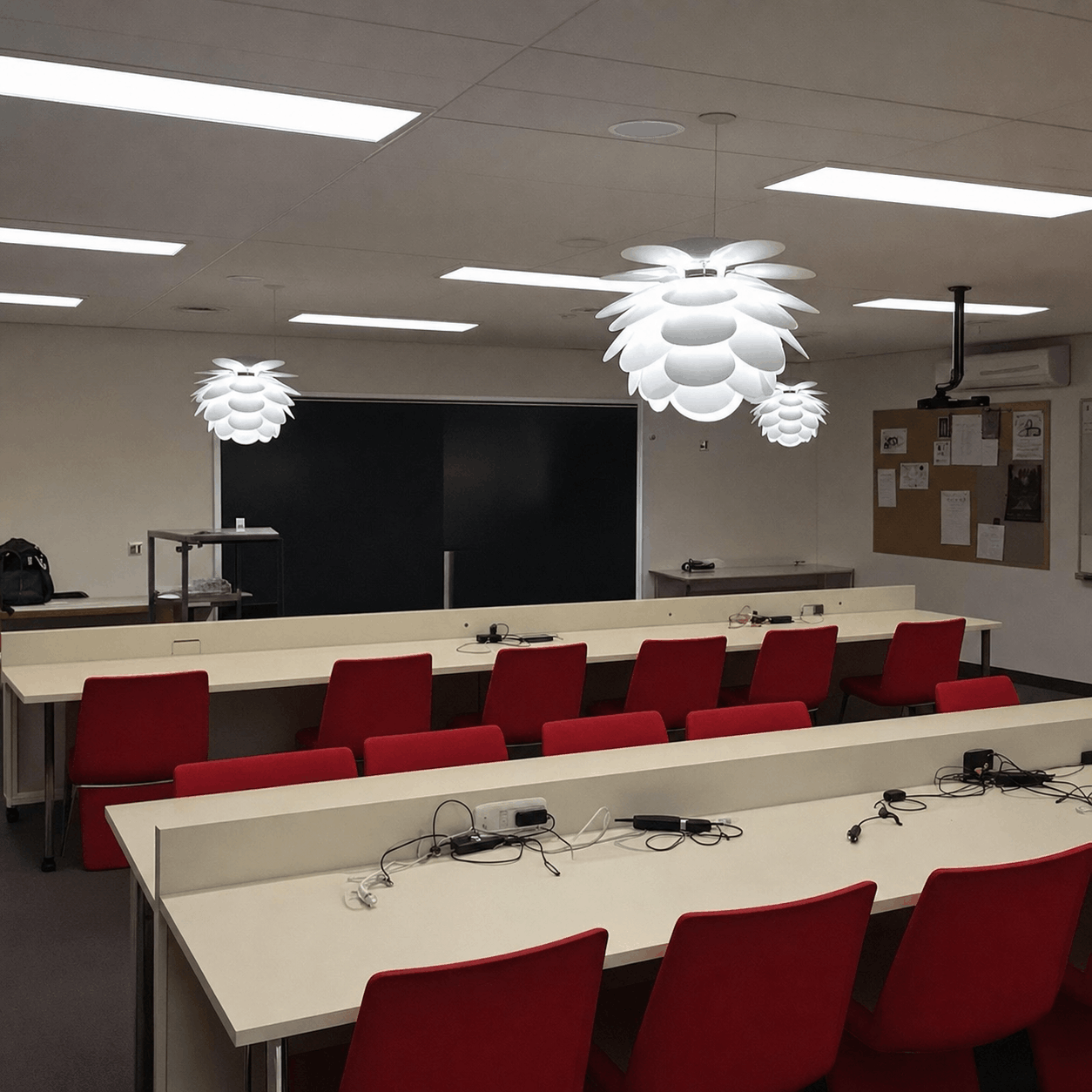 A classroom with red chairs and white tables, featuring modern white pendant lights. The room is well-lit, with a large blackboard and bulletin board.