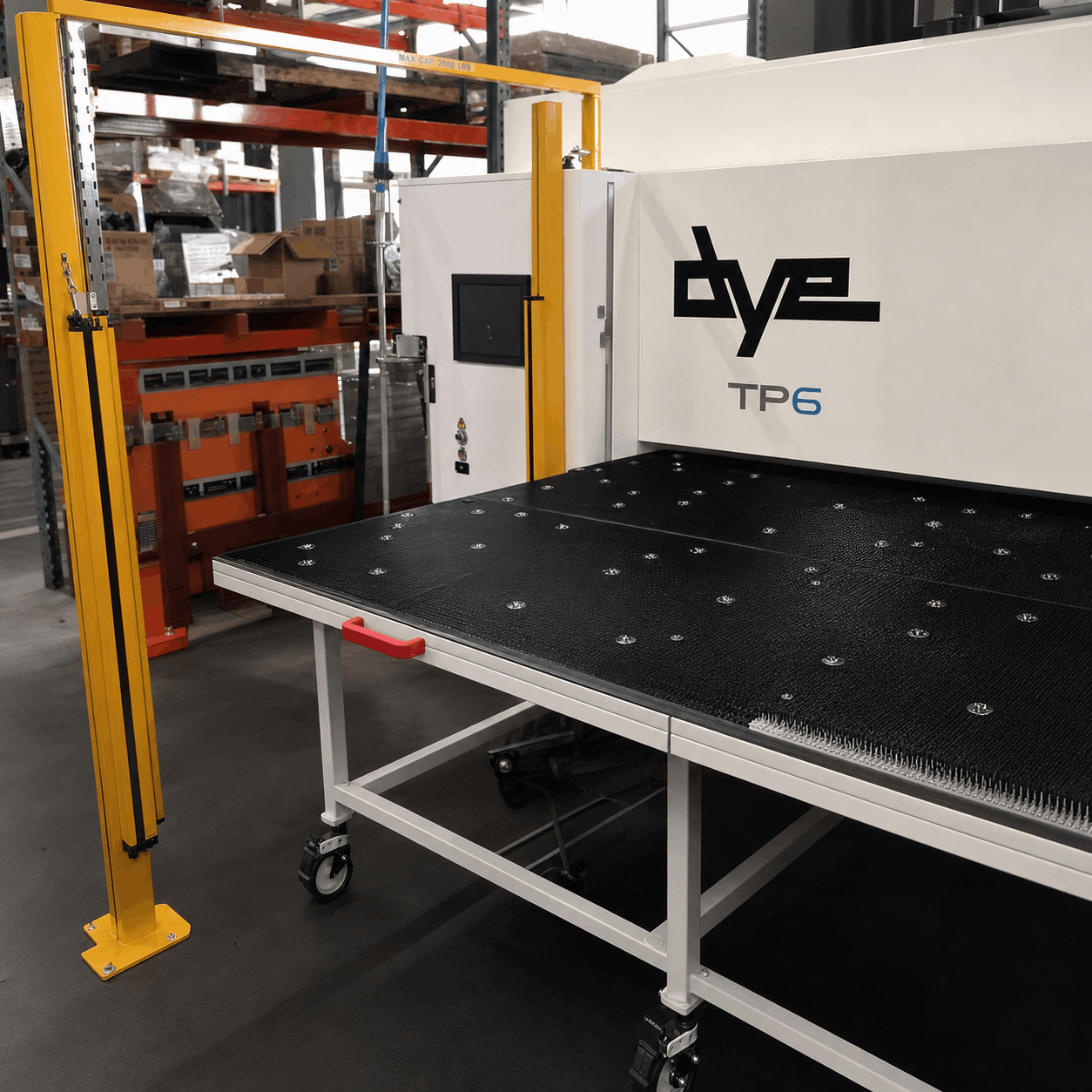 Industrial machine in a warehouse setting. It has a black, textured worktable with metal knobs, yellow safety frame, and shelves with boxes in the background.