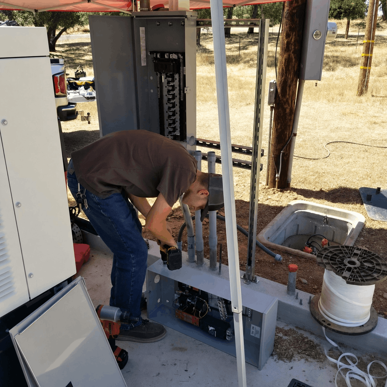 A person works on electrical equipment outdoors under a canopy, surrounded by tools and cables, suggesting a focused and industrious atmosphere.