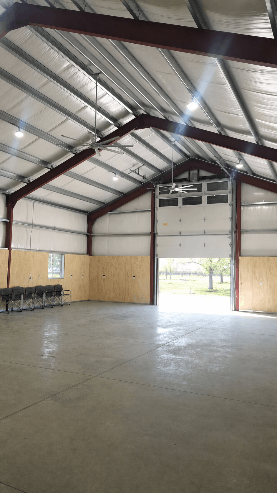 Spacious industrial warehouse interior with high metal ceiling, large open garage door, and natural light. Rows of empty chairs line one wall.