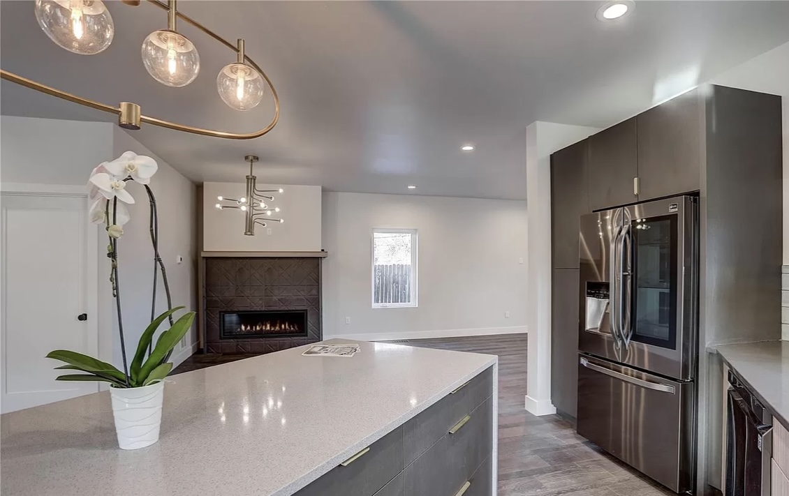 Modern kitchen interior with sleek gray cabinetry, stainless steel fridge, large island with orchid centerpiece, and a stylish chandelier. Cozy fireplace in the background.