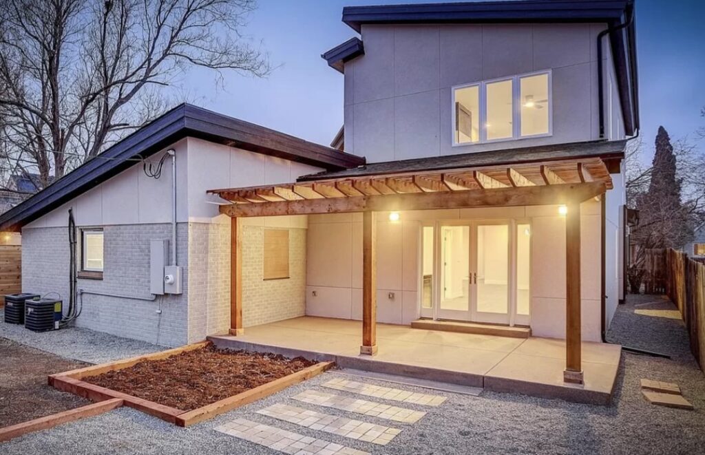 Modern house exterior at dusk with warm lights illuminating a covered patio. Features include a wood pergola and spacious windows, creating an inviting atmosphere.