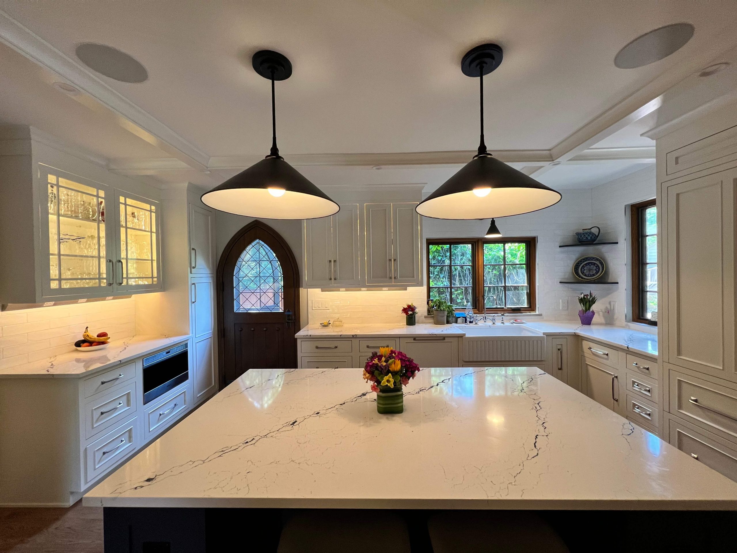 Bright kitchen with marble countertops, dark pendant lights, and a wooden arched door. A vase of flowers sits on the island, creating a cozy ambience.