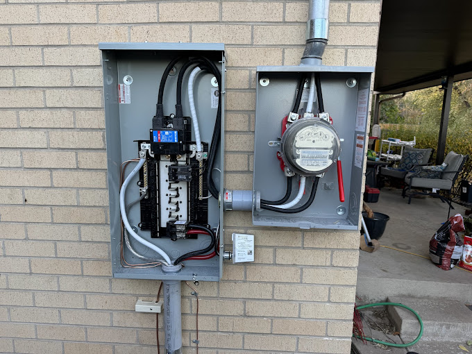 Electrical panel mounted on a brick wall with visible wiring and a connected meter. The setup is outdoors, near a covered patio with some chairs.