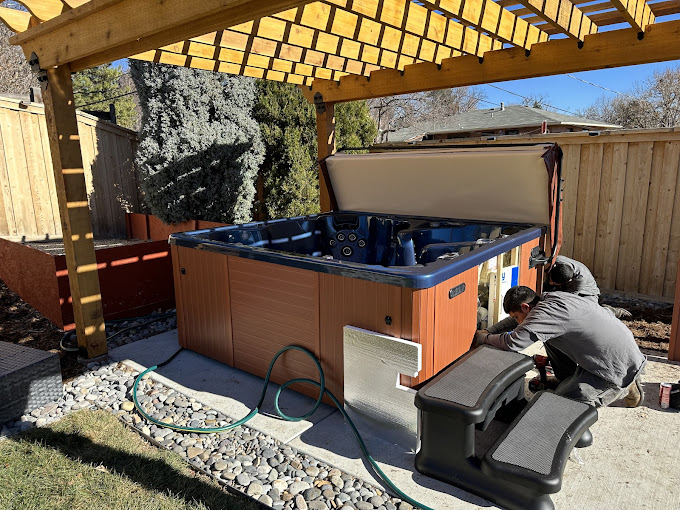 A wooden hot tub with an open lid sits under a pergola in a backyard. Two people work on its side, surrounded by pebbles and greenery on a sunny day.