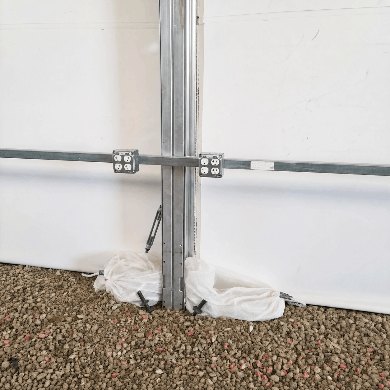 Metal brackets securing a vertical metal beam to a white wall, surrounded by gravel ground and some white protective material at the base.