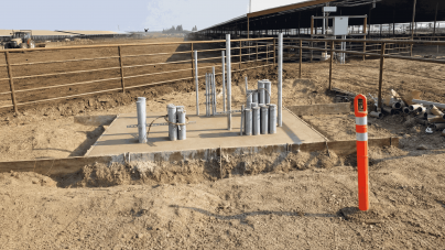 Construction site with metal pipes protruding from a concrete foundation, surrounded by dirt. A bright orange safety bollard stands nearby.