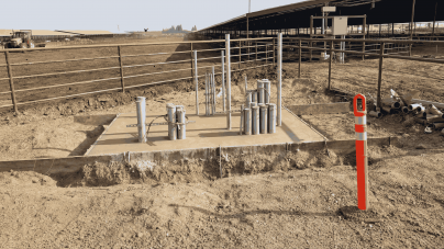 Construction site with metal pipes protruding from a concrete foundation, surrounded by dirt. A bright orange safety bollard stands nearby.