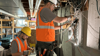 Two electricians work in a dimly lit basement. One stands at an open electrical panel with wiring exposed, while the other inspects tools.
