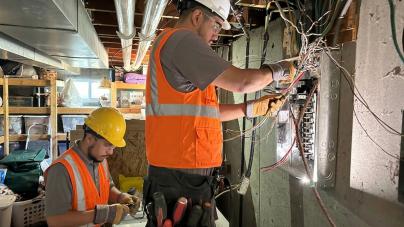 Two electricians work in a dimly lit basement. One stands at an open electrical panel with wiring exposed, while the other inspects tools.