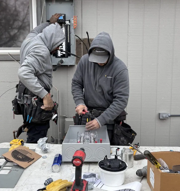 Two electricians in gray hoodies work on an outdoor electrical panel. One is adjusting wires while the other uses a screwdriver at a workbench. Tools and equipment are scattered on the table, conveying a focused and industrious atmosphere.