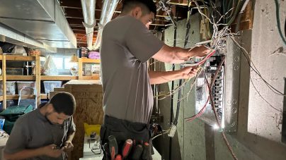 Two electricians work in a dimly lit basement. One stands at an open electrical panel with wiring exposed, while the other inspects tools.